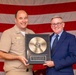 A U.S. Navy servicemember in tan/khaki uniform and a civilian man in a navy blue suit are jointly holding a commemorative gold record plaque in front of an American flag. The plaque honors Dennis P. Connell for his service from April 5, 2005 to August 31, 2026, with the inscription 'Fair Winds, Following Seas, and Louder Speakers.' This appears to be a military retirement or service recognition ceremony.