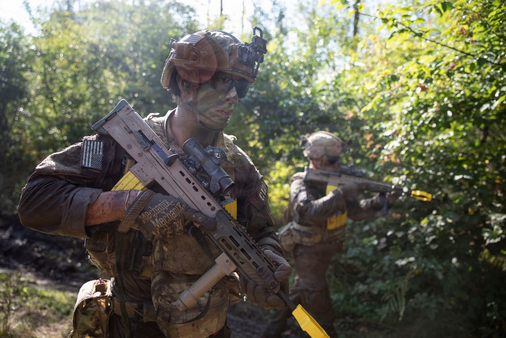 Two military personnel in full combat gear are conducting what appears to be a training exercise in a wooded environment. The foreground soldier wears camouflage face paint, a tactical helmet with night-vision mount, and carries a modern bullpup-style rifle with yellow training indicators. Both soldiers are equipped with US flag patches and Multicam/OCP pattern uniforms.