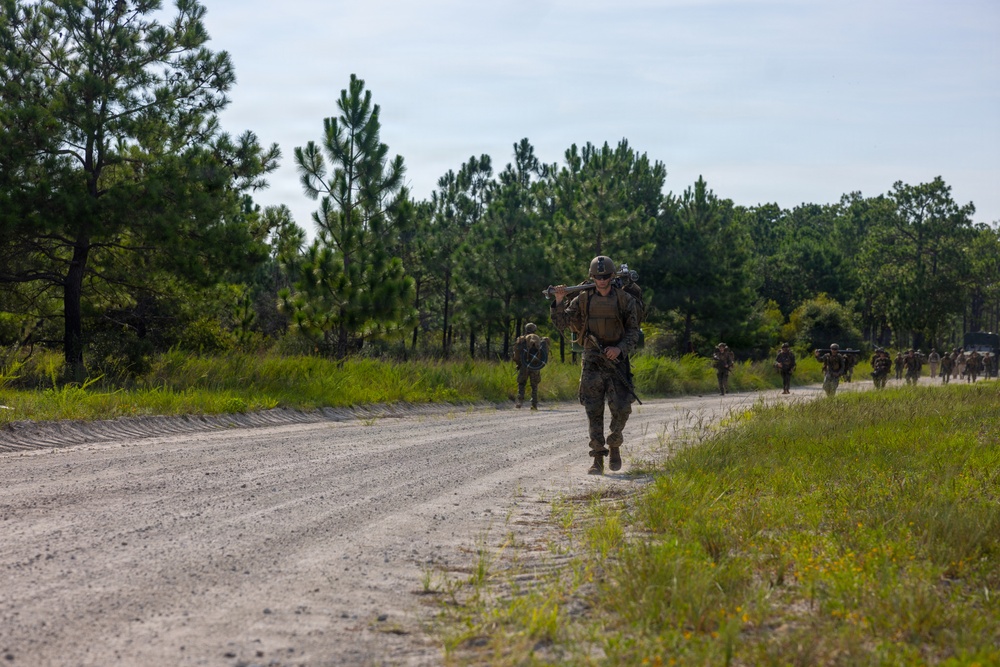 A column of military personnel in combat gear and camouflage uniforms conducts a road march along a sandy gravel road through a pine tree-lined training area. The lead soldier carries a rifle across their shoulders and wears a full pack and helmet. The scene appears to be a military training exercise, likely at a U.S. military installation based on the terrain and vegetation.