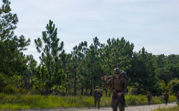 A column of military personnel in combat gear and camouflage uniforms conducts a road march along a sandy gravel road through a pine tree-lined training area. The lead soldier carries a rifle across their shoulders and wears a full pack and helmet. The scene appears to be a military training exercise, likely at a U.S. military installation based on the terrain and vegetation.