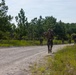 A column of military personnel in combat gear and camouflage uniforms conducts a road march along a sandy gravel road through a pine tree-lined training area. The lead soldier carries a rifle across their shoulders and wears a full pack and helmet. The scene appears to be a military training exercise, likely at a U.S. military installation based on the terrain and vegetation.