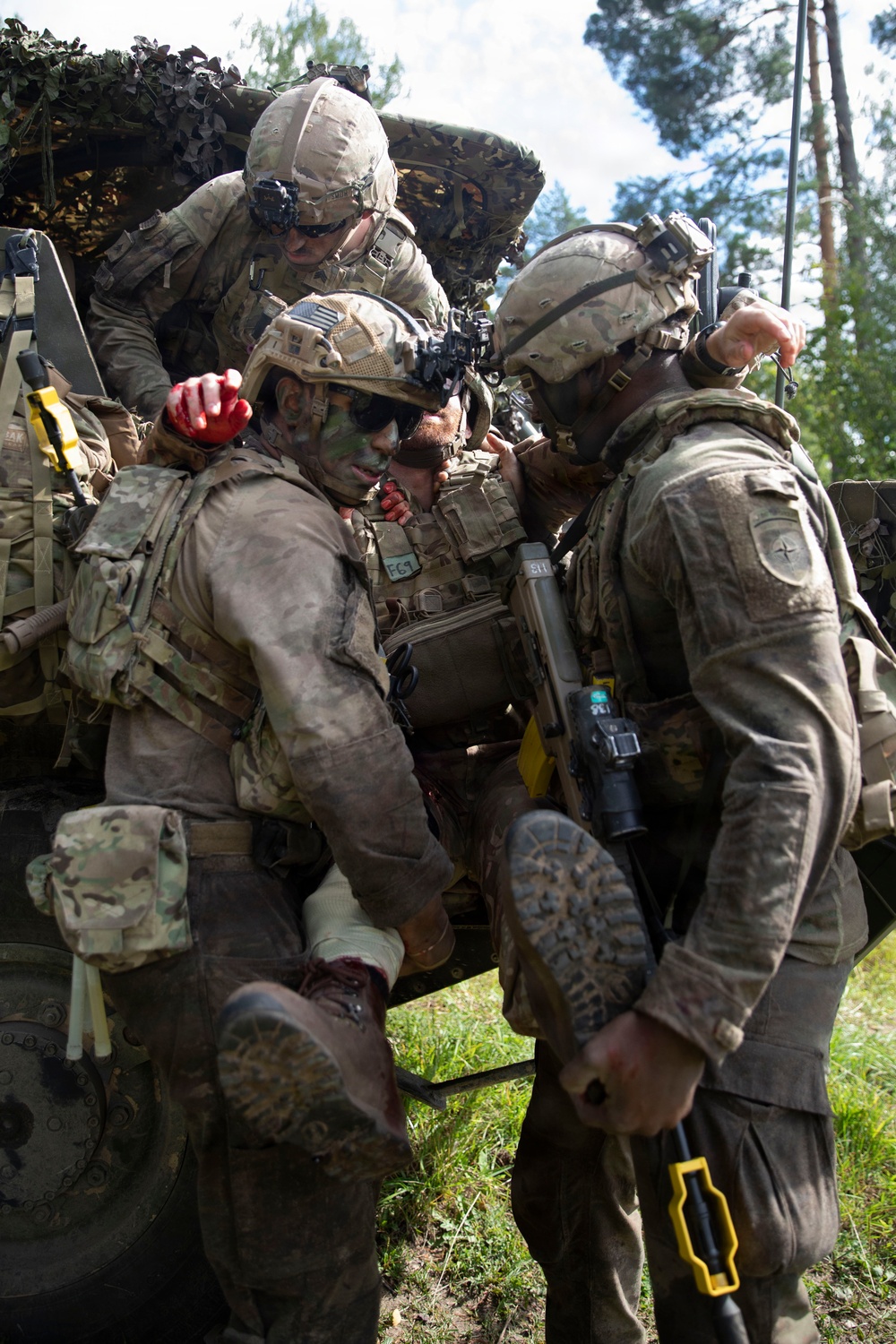 Multiple soldiers in full combat gear and MultiCam/OCP uniforms are conducting a casualty evacuation (CASEVAC) drill, extracting a simulated wounded soldier from a military vehicle. The soldiers display face camouflage paint, tactical helmets with night-vision mounts, and body armor. Simulated blood (moulage) is visible on the casualty's hands and legs, indicating this is a training exercise.