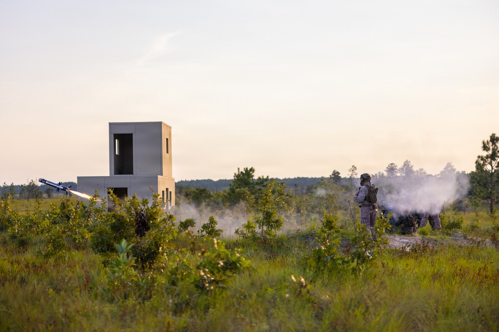 A military training exercise is underway in an open field environment. A soldier in combat gear is visible on the right side as a missile or rocket is being launched from near a concrete training structure on the left. Smoke and exhaust plume are clearly visible from the launch event.