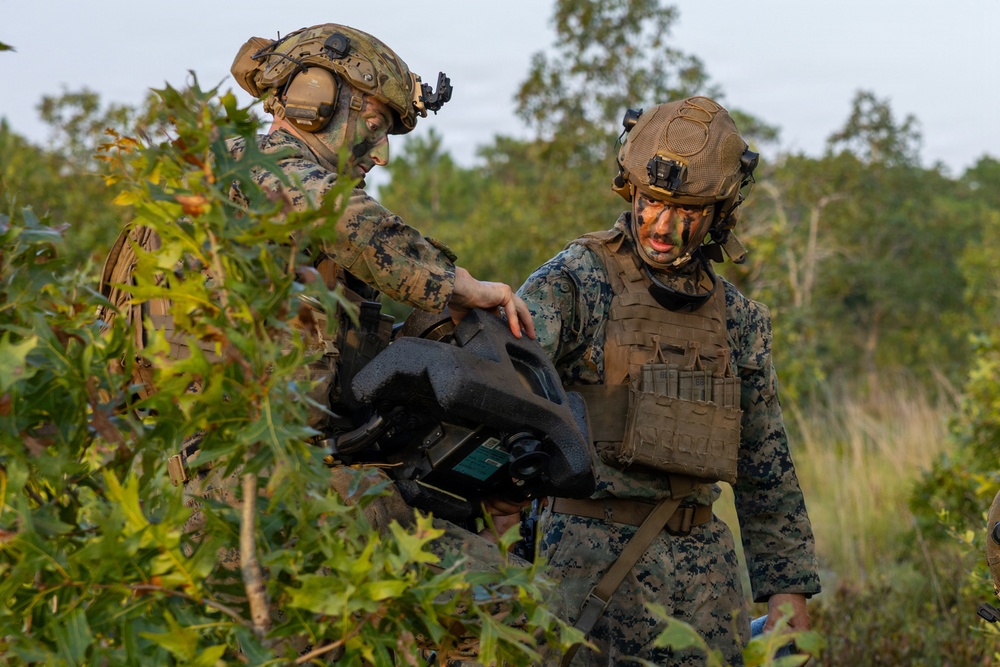 Two military personnel in MARPAT (Marine pattern) camouflage uniforms are handling what appears to be a drone or unmanned aerial vehicle (UAV) case in a wooded/brushy outdoor environment. Both individuals are wearing tactical gear including plate carriers, combat helmets with night vision mounts, and communications headsets. Their faces are painted with camouflage face paint, suggesting a field training exercise or operational deployment.