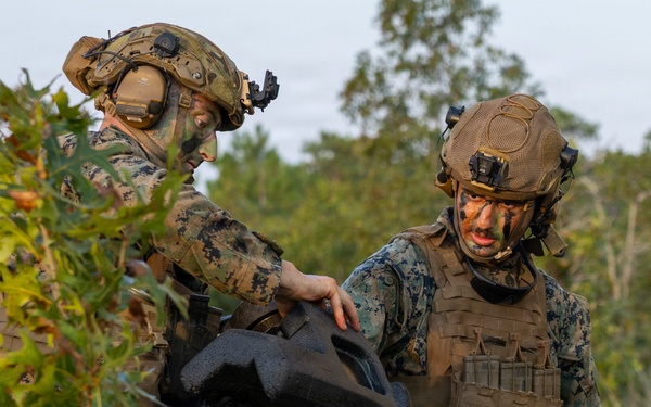 Two military personnel in MARPAT (Marine pattern) camouflage uniforms are handling what appears to be a drone or unmanned aerial vehicle (UAV) case in a wooded/brushy outdoor environment. Both individuals are wearing tactical gear including plate carriers, combat helmets with night vision mounts, and communications headsets. Their faces are painted with camouflage face paint, suggesting a field training exercise or operational deployment.