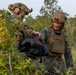 Two military personnel in MARPAT (Marine pattern) camouflage uniforms are handling what appears to be a drone or unmanned aerial vehicle (UAV) case in a wooded/brushy outdoor environment. Both individuals are wearing tactical gear including plate carriers, combat helmets with night vision mounts, and communications headsets. Their faces are painted with camouflage face paint, suggesting a field training exercise or operational deployment.