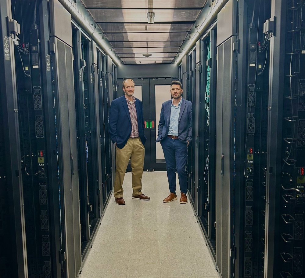 Two professionally dressed men are standing in the aisle of a data center, flanked by rows of black server racks on both sides. The setting appears to be a modern, well-maintained server room with overhead cable management and cooling infrastructure. The image appears to be a posed professional or promotional photograph.