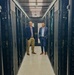 Two professionally dressed men are standing in the aisle of a data center, flanked by rows of black server racks on both sides. The setting appears to be a modern, well-maintained server room with overhead cable management and cooling infrastructure. The image appears to be a posed professional or promotional photograph.