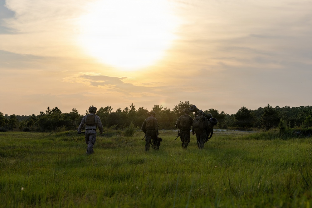 A group of approximately five military personnel in full combat gear are walking away from the camera across an open grassy field toward a tree line. The image is captured at either sunrise or sunset, creating a dramatic backlit silhouette effect. The soldiers appear to be on patrol or returning from a field exercise.