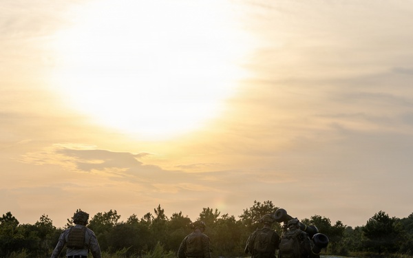 A group of approximately five military personnel in full combat gear are walking away from the camera across an open grassy field toward a tree line. The image is captured at either sunrise or sunset, creating a dramatic backlit silhouette effect. The soldiers appear to be on patrol or returning from a field exercise.