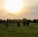 A group of approximately five military personnel in full combat gear are walking away from the camera across an open grassy field toward a tree line. The image is captured at either sunrise or sunset, creating a dramatic backlit silhouette effect. The soldiers appear to be on patrol or returning from a field exercise.