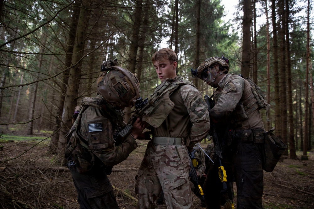 Three individuals in military camouflage uniforms are gathered in a forested area, appearing to conduct a search or detainment procedure on the central figure. The soldier on the left displays a US flag patch on their shoulder and is bent forward examining or restraining the middle person. The setting and equipment suggest a military training exercise rather than active combat.