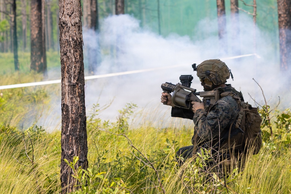 A military service member in full combat gear is positioned in a wooded area, aiming a weapon system toward a smoke-filled clearing. The individual is wearing digital camouflage (MARPAT pattern), a tactical helmet, and a backpack, suggesting U.S. Marine Corps affiliation. White smoke is visible in the background, consistent with a training exercise environment.