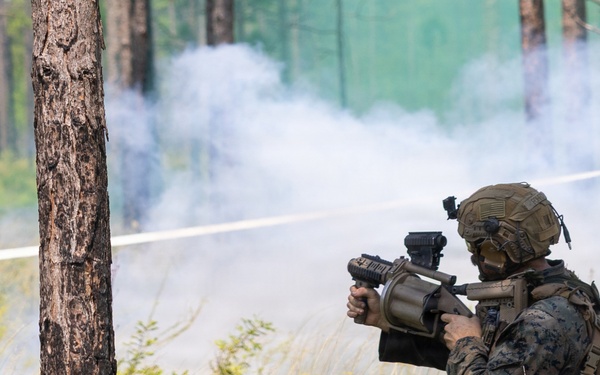 A military service member in full combat gear is positioned in a wooded area, aiming a weapon system toward a smoke-filled clearing. The individual is wearing digital camouflage (MARPAT pattern), a tactical helmet, and a backpack, suggesting U.S. Marine Corps affiliation. White smoke is visible in the background, consistent with a training exercise environment.