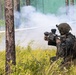A military service member in full combat gear is positioned in a wooded area, aiming a weapon system toward a smoke-filled clearing. The individual is wearing digital camouflage (MARPAT pattern), a tactical helmet, and a backpack, suggesting U.S. Marine Corps affiliation. White smoke is visible in the background, consistent with a training exercise environment.