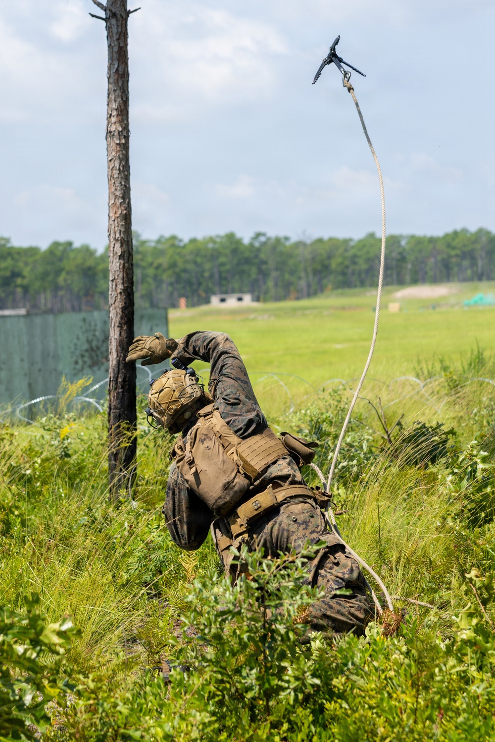 A military service member in MARPAT camouflage uniform is lying in vegetation and throwing a grappling hook attached to a rope into the air. The action appears to be part of a training exercise, likely aimed at breaching a wire obstacle or scaling a barrier. The setting is an open field training area with a treeline and structures visible in the background.