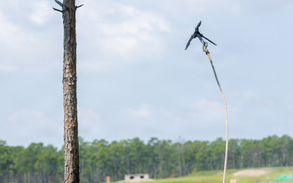 A military service member in MARPAT camouflage uniform is lying in vegetation and throwing a grappling hook attached to a rope into the air. The action appears to be part of a training exercise, likely aimed at breaching a wire obstacle or scaling a barrier. The setting is an open field training area with a treeline and structures visible in the background.