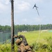 A military service member in MARPAT camouflage uniform is lying in vegetation and throwing a grappling hook attached to a rope into the air. The action appears to be part of a training exercise, likely aimed at breaching a wire obstacle or scaling a barrier. The setting is an open field training area with a treeline and structures visible in the background.