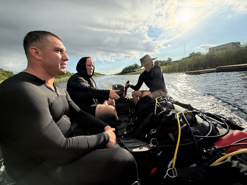 Three individuals are traveling in a small inflatable boat along a narrow waterway, likely a canal or river. Two of the men are wearing black wetsuits or dive skins, while a third person in the rear operates the outboard motor while wearing a boonie hat. The boat is loaded with scuba diving equipment including tanks, BCDs, and hoses, suggesting a dive operation or training exercise.