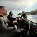 Three individuals are traveling in a small inflatable boat along a narrow waterway, likely a canal or river. Two of the men are wearing black wetsuits or dive skins, while a third person in the rear operates the outboard motor while wearing a boonie hat. The boat is loaded with scuba diving equipment including tanks, BCDs, and hoses, suggesting a dive operation or training exercise.