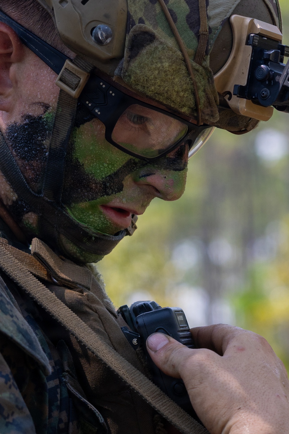 A close-up photograph of a military service member wearing camouflage face paint, a tactical helmet with accessories, and protective eyewear. The individual is looking downward at a small black radio or communication device being held at chest level. The image appears to depict a field training exercise or operational environment, evidenced by the wet skin and full tactical gear.