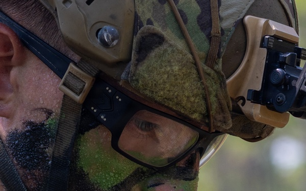 A close-up photograph of a military service member wearing camouflage face paint, a tactical helmet with accessories, and protective eyewear. The individual is looking downward at a small black radio or communication device being held at chest level. The image appears to depict a field training exercise or operational environment, evidenced by the wet skin and full tactical gear.