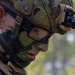 A close-up photograph of a military service member wearing camouflage face paint, a tactical helmet with accessories, and protective eyewear. The individual is looking downward at a small black radio or communication device being held at chest level. The image appears to depict a field training exercise or operational environment, evidenced by the wet skin and full tactical gear.