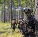 A group of military personnel in full combat gear patrol through a pine forest during what appears to be a training exercise. The foreground soldier is equipped with camouflage face paint, a tactical helmet with night vision mount, radio antenna, rifle, and body armor with pouches. Additional soldiers are visible in the background moving through the wooded terrain.