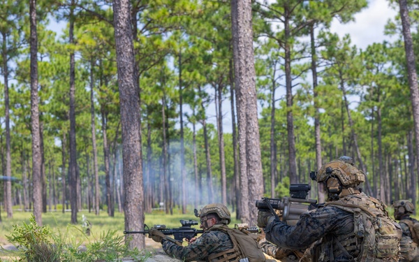 Military personnel, appearing to be U.S. Marines based on their MARPAT camouflage uniforms, are conducting a field training exercise in a pine forest environment. Two foreground personnel are in firing positions behind a sandy berm, with visible smoke indicating live or blank fire discharge. Additional personnel are visible in the background, suggesting a coordinated tactical drill.