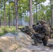 Military personnel, appearing to be U.S. Marines based on their MARPAT camouflage uniforms, are conducting a field training exercise in a pine forest environment. Two foreground personnel are in firing positions behind a sandy berm, with visible smoke indicating live or blank fire discharge. Additional personnel are visible in the background, suggesting a coordinated tactical drill.