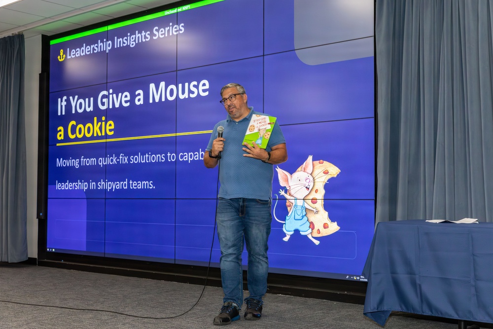 A man is presenting at a 'Leadership Insights Series' event, holding a microphone and the children's book 'If You Give a Mouse a Cookie.' The presentation slide behind him uses the book as a metaphor for 'Moving from quick-fix solutions to capable leadership in shipyard teams.' The setting appears to be a professional conference or training room with a large video wall display.