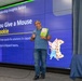 A man is presenting at a 'Leadership Insights Series' event, holding a microphone and the children's book 'If You Give a Mouse a Cookie.' The presentation slide behind him uses the book as a metaphor for 'Moving from quick-fix solutions to capable leadership in shipyard teams.' The setting appears to be a professional conference or training room with a large video wall display.