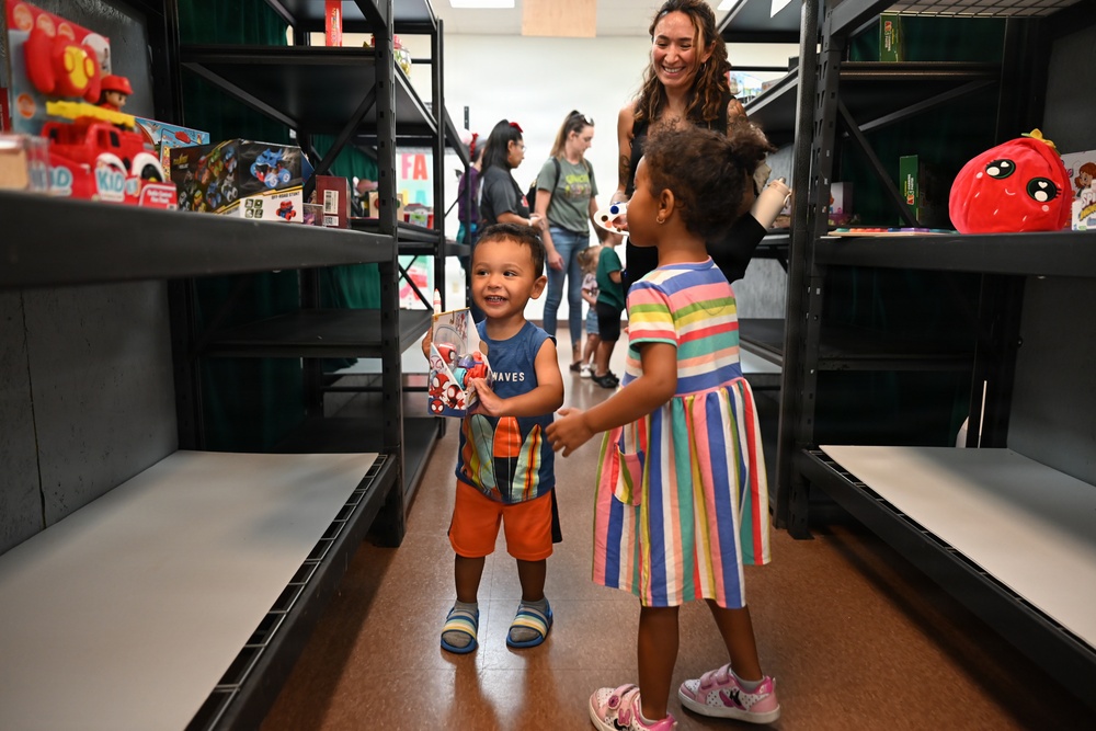 Two young children interact in an aisle between dark metal shelving units in what appears to be a toy distribution or charity event. A smiling adult woman watches over them from behind, while other people are visible in the background. The boy is holding a packaged toy while facing a girl in a colorful striped dress.