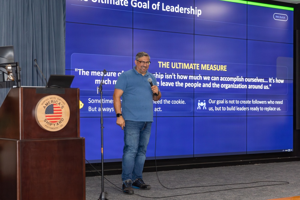 A male speaker is presenting a leadership seminar at what appears to be a Naval facility, standing in front of a large video wall display showing slide 10 of 10 titled 'The Ultimate Goal of Leadership.' He is holding a microphone and standing near a wooden podium bearing the 'America's Shipyard' emblem, identified as Norfolk Naval Shipyard, Portsmouth, VA. The presentation slide features a section called 'The Ultimate Measure' with quotes about leadership philosophy.