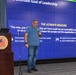 A male speaker is presenting a leadership seminar at what appears to be a Naval facility, standing in front of a large video wall display showing slide 10 of 10 titled 'The Ultimate Goal of Leadership.' He is holding a microphone and standing near a wooden podium bearing the 'America's Shipyard' emblem, identified as Norfolk Naval Shipyard, Portsmouth, VA. The presentation slide features a section called 'The Ultimate Measure' with quotes about leadership philosophy.