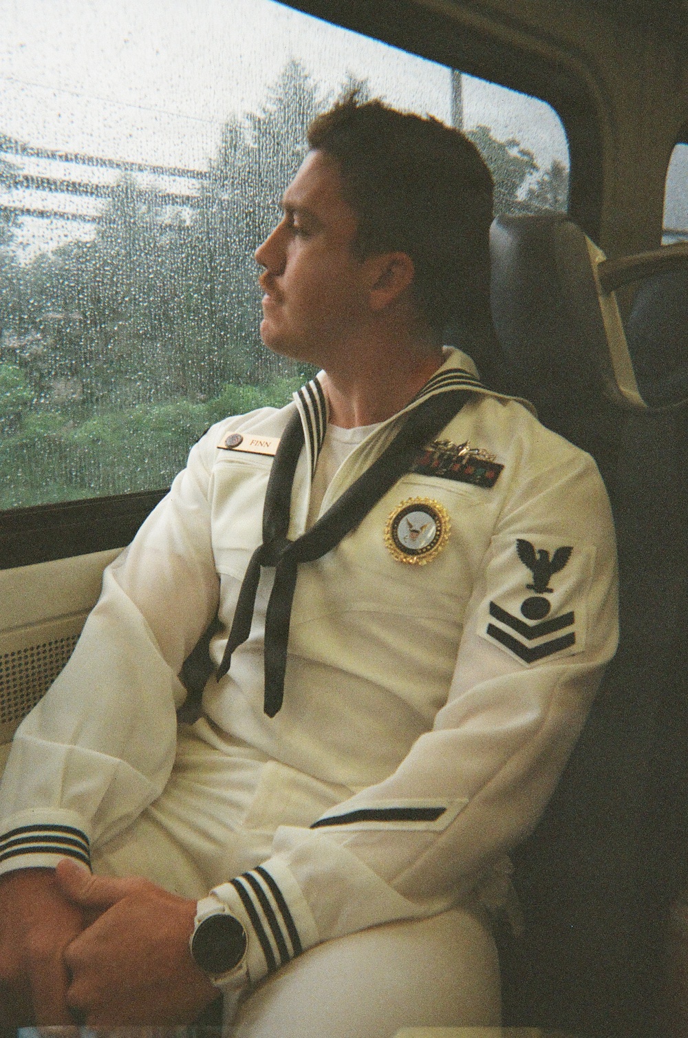 A young male U.S. Navy sailor in dress white uniform is seated on what appears to be a train or bus, gazing out a rain-streaked window. He is wearing a formal Service Dress White uniform with visible rank insignia, ribbons, a name tag reading 'FINN,' and a black neckerchief. The photo has a film photography aesthetic with warm tones.