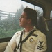 A young male U.S. Navy sailor in dress white uniform is seated on what appears to be a train or bus, gazing out a rain-streaked window. He is wearing a formal Service Dress White uniform with visible rank insignia, ribbons, a name tag reading 'FINN,' and a black neckerchief. The photo has a film photography aesthetic with warm tones.