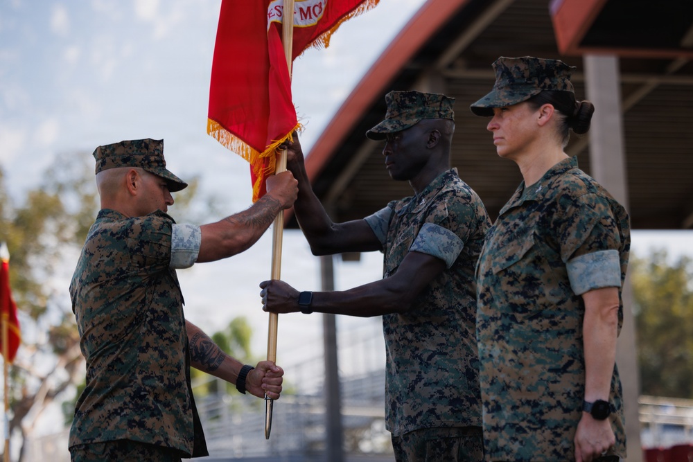 Three U.S. Marines in MARPAT camouflage uniforms are participating in a formal military ceremony involving the transfer of a red guidon or unit colors. Two Marines are actively exchanging the flag staff, which is a traditional element of a change of command or activation/deactivation ceremony. The event appears to be taking place outdoors at a military installation, likely in a warm climate.
