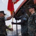 Three U.S. Marines in MARPAT camouflage uniforms are participating in a formal military ceremony involving the transfer of a red guidon or unit colors. Two Marines are actively exchanging the flag staff, which is a traditional element of a change of command or activation/deactivation ceremony. The event appears to be taking place outdoors at a military installation, likely in a warm climate.
