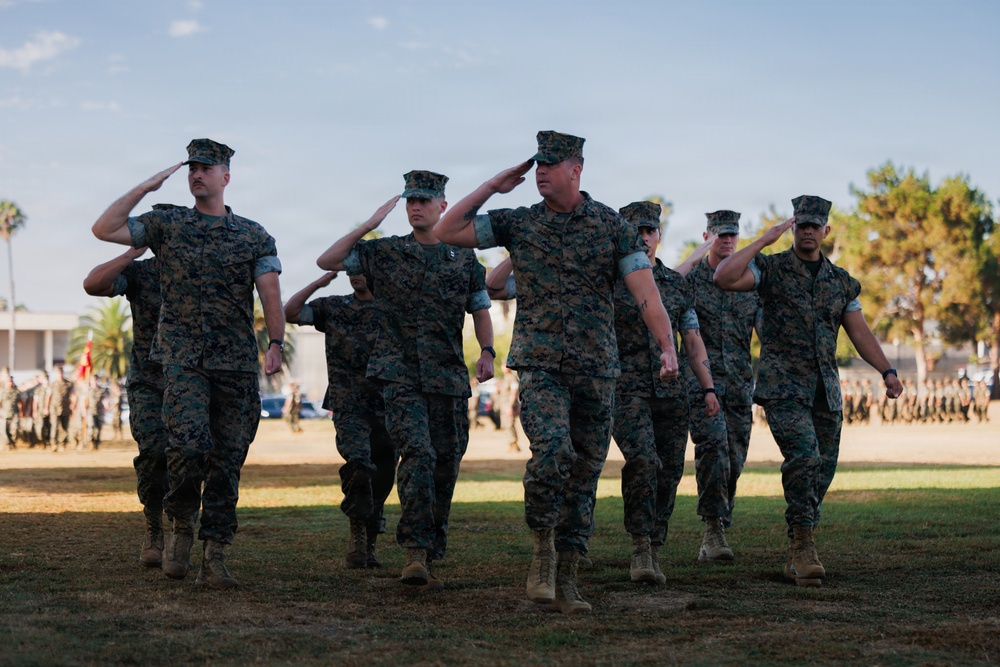 A group of U.S. Marines in woodland MARPAT camouflage uniforms are marching in formation while rendering a salute during what appears to be a formal military ceremony or parade. The event is taking place on a grass field with a large formation of troops visible in the background. The setting appears to be a military installation, likely on the West Coast based on the palm trees and vegetation.