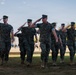 A group of U.S. Marines in woodland MARPAT camouflage uniforms are marching in formation while rendering a salute during what appears to be a formal military ceremony or parade. The event is taking place on a grass field with a large formation of troops visible in the background. The setting appears to be a military installation, likely on the West Coast based on the palm trees and vegetation.