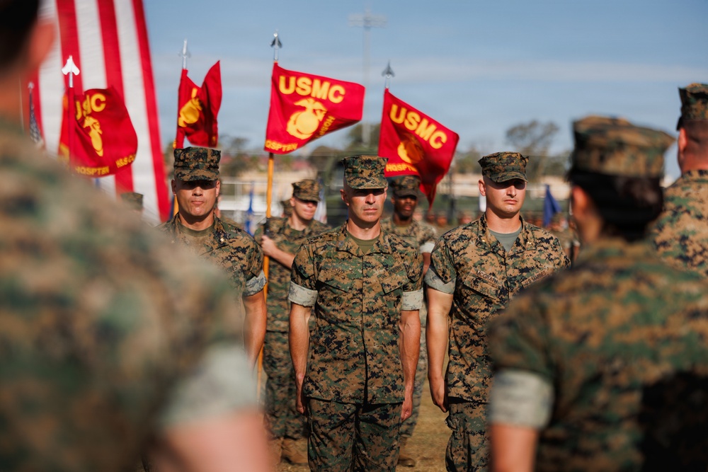 A formal United States Marine Corps (USMC) military ceremony is taking place outdoors, likely on a parade deck or drill field. Multiple Marines in MARPAT woodland camouflage uniforms are standing at attention while color guard members carry USMC guidons and an American flag. The scene appears to be a formal military formation event such as a change of command, promotion, or unit ceremony.