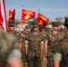 A formal United States Marine Corps (USMC) military ceremony is taking place outdoors, likely on a parade deck or drill field. Multiple Marines in MARPAT woodland camouflage uniforms are standing at attention while color guard members carry USMC guidons and an American flag. The scene appears to be a formal military formation event such as a change of command, promotion, or unit ceremony.
