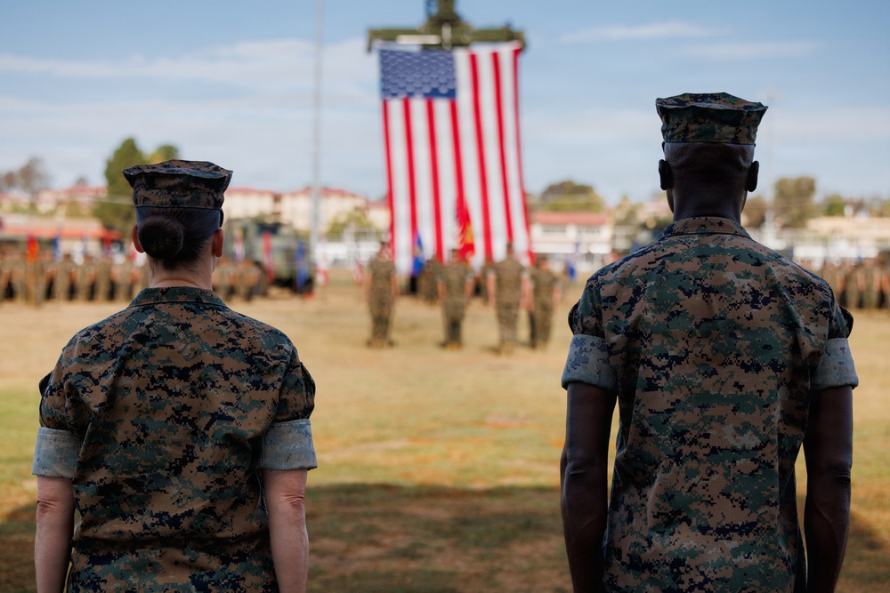 Two U.S. Marines in digital camouflage uniforms (MARPAT) stand at attention facing a large American flag during a formal military ceremony. A formation of additional Marines is visible in the background on a parade field. The setting appears to be a military installation, likely a change of command or similar ceremonial event.