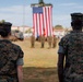 Two U.S. Marines in digital camouflage uniforms (MARPAT) stand at attention facing a large American flag during a formal military ceremony. A formation of additional Marines is visible in the background on a parade field. The setting appears to be a military installation, likely a change of command or similar ceremonial event.