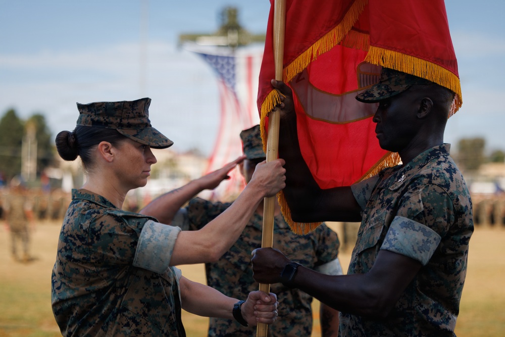 Two U.S. Marine Corps personnel in MARPAT digital camouflage uniforms are participating in a formal military ceremony involving the transfer of a red guidon or unit colors with gold fringe. The ceremony appears to be a change of command or activation/deactivation event taking place on a parade field with formations of Marines visible in the background. An American flag is visible in the blurred background.