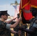 Two U.S. Marine Corps personnel in MARPAT digital camouflage uniforms are participating in a formal military ceremony involving the transfer of a red guidon or unit colors with gold fringe. The ceremony appears to be a change of command or activation/deactivation event taking place on a parade field with formations of Marines visible in the background. An American flag is visible in the blurred background.