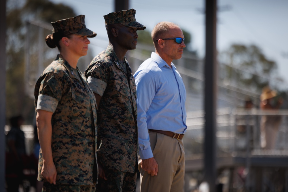 Three individuals stand at attention in an outdoor military setting. Two are wearing U.S. Marine Corps MARPAT camouflage uniforms with covers (caps), while the third is a civilian male in a light blue dress shirt and khaki pants with sunglasses. They appear to be observing a formal military event or ceremony.