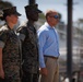 Three individuals stand at attention in an outdoor military setting. Two are wearing U.S. Marine Corps MARPAT camouflage uniforms with covers (caps), while the third is a civilian male in a light blue dress shirt and khaki pants with sunglasses. They appear to be observing a formal military event or ceremony.