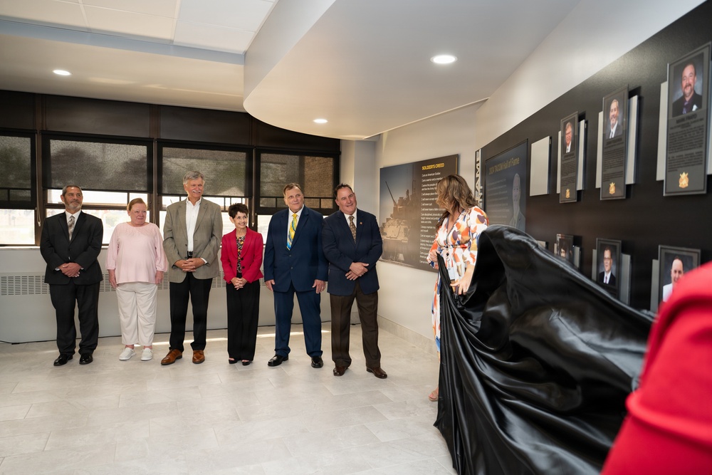A formal unveiling ceremony is taking place in what appears to be a hall of fame or memorial corridor. A woman in a colorful dress is pulling back a black drape to reveal framed portraits and plaques mounted on a dark wall, while six attendees stand and observe. The wall displays what appears to be a 'Soldier's Creed' panel and multiple framed photographs with biographical plaques, suggesting a military or institutional recognition display.
